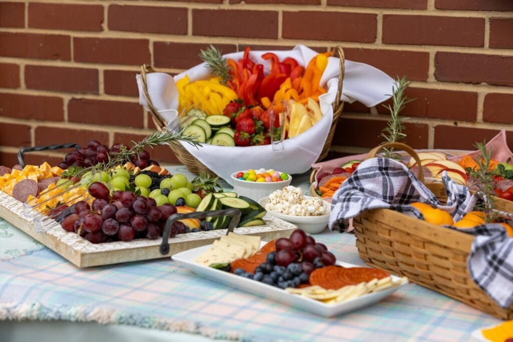Fresh fruit and vegetable platter at The Enchanted Event C, featuring grapes, strawberries, cucumbers, peppers, cheese, crackers, and assorted snacks, arranged on a picnic table against a brick wall.