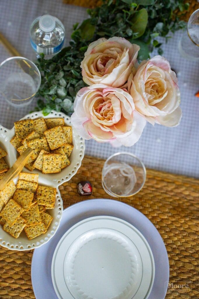 Lavender and cream-colored roses arranged in a bouquet on a table decorated for The Enchanted Event C, with snacks, wine glasses, and greenery.